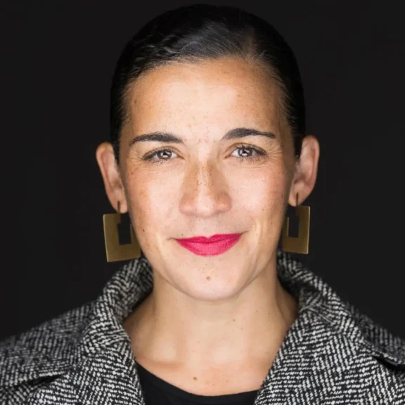 A woman with dark hair, wearing geometric earrings and a patterned jacket, faces the camera against a black background.
