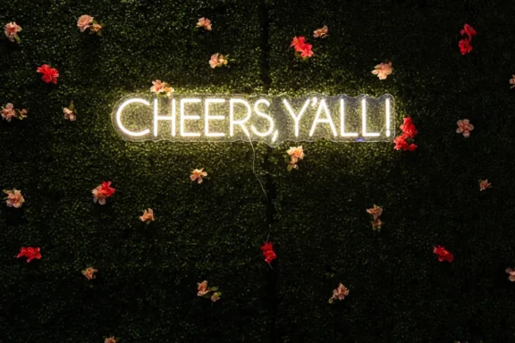 Neon sign reading "CHEERS Y'ALL!" mounted on a green wall with scattered pink and red artificial flowers.
