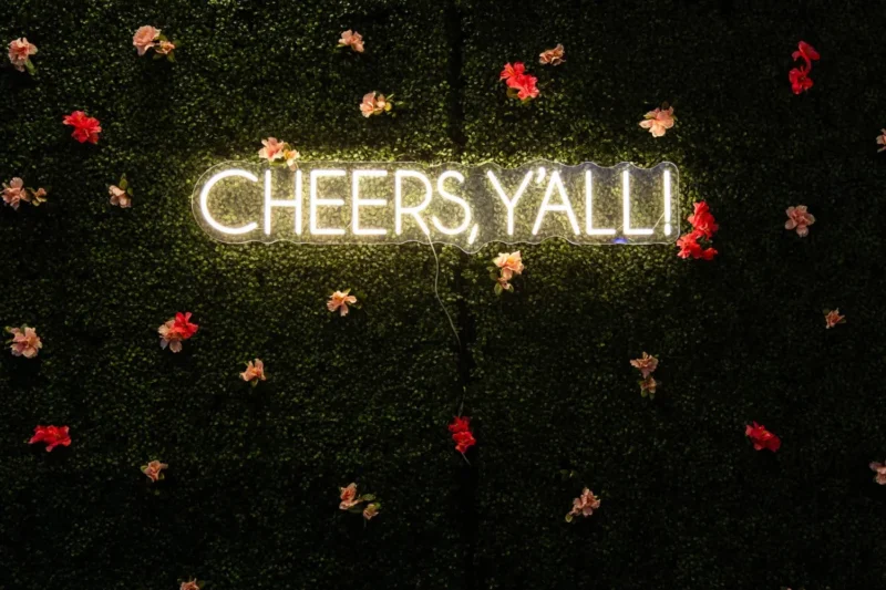 Neon sign reading "CHEERS Y'ALL!" mounted on a green wall with scattered pink and red artificial flowers.