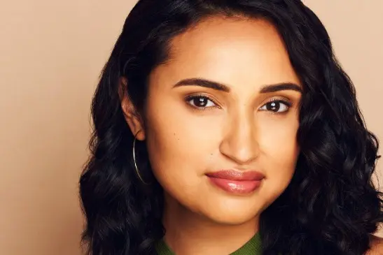 A woman with medium skin tone, long wavy dark hair, and hoop earrings looks at the camera against a plain beige background.