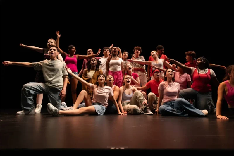 A diverse group of performers on stage, some kneeling and others standing, all reaching and singing passionately under stage lighting.