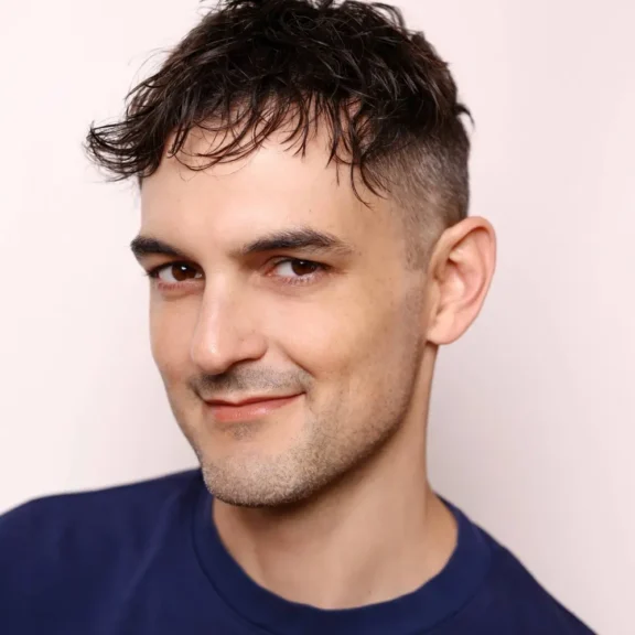 A man with short, dark, wavy hair and a trimmed beard is wearing a navy blue shirt and smiling slightly at the camera.