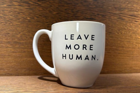 A white ceramic mug with the words "LEAVE MORE HUMAN." printed in black uppercase letters, placed on a wooden surface with a wooden background.