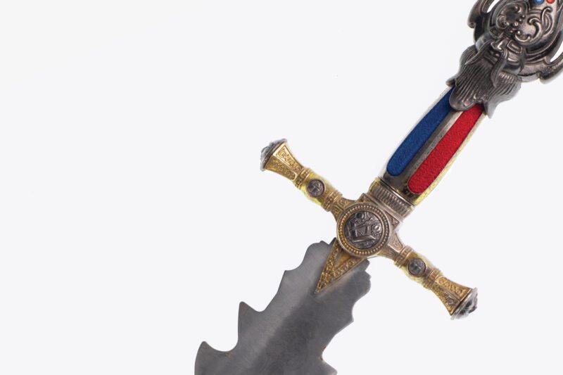 Close-up of an ornate sword with a jagged blade, gold and silver hilt, red and blue grip, and decorative details on the crossguard and pommel, set against a white background.