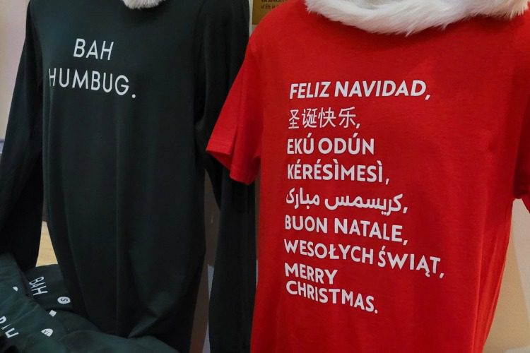 Two t-shirts: one black with "BAH HUMBUG." in white text, and one red with "Merry Christmas" written in multiple languages in white text.
