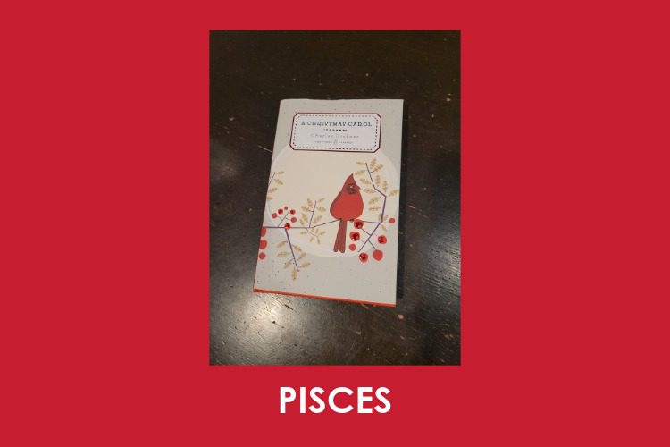 A book titled "A Christmas Carol" with a red bird illustration on the cover sits on a dark wooden surface. The word "PISCES" is written below on a red background.
