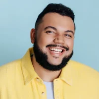 A man with a short beard and dark hair smiles at the camera, wearing a yellow jacket over a white shirt against a light blue background.