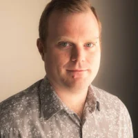 A man with light skin and short hair wearing a patterned button-up shirt stands in front of a neutral background, looking at the camera.