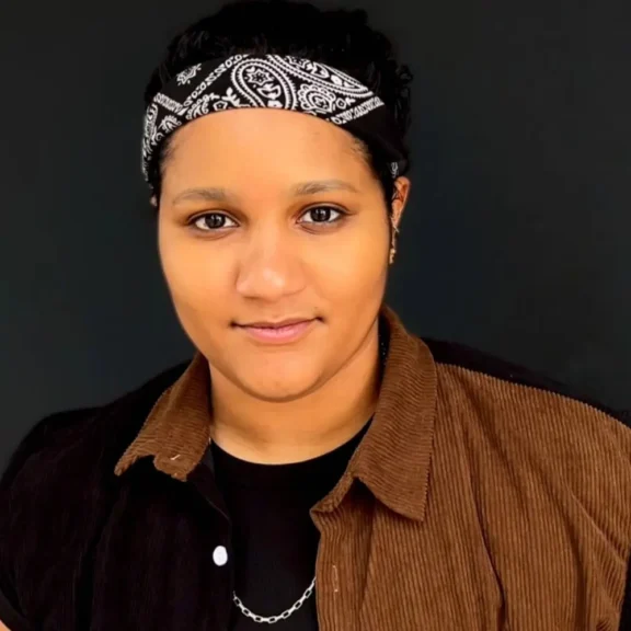 A person with short dark hair wears a black and white bandana, a brown corduroy shirt over a black top, and a chain necklace, posing against a plain black background.