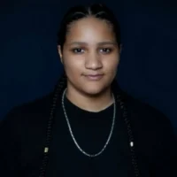 A person with long braids, wearing a black shirt, black jacket, and silver chain necklace, poses against a dark background.