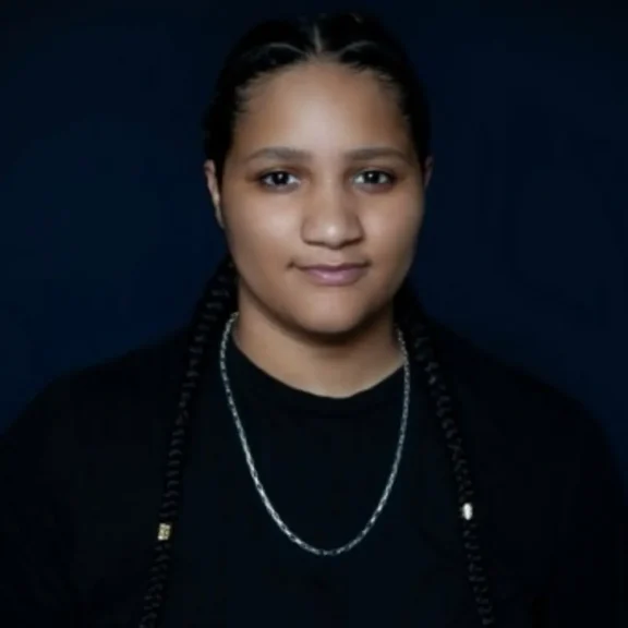 A person with long braids, wearing a black shirt, black jacket, and silver chain necklace, poses against a dark background.