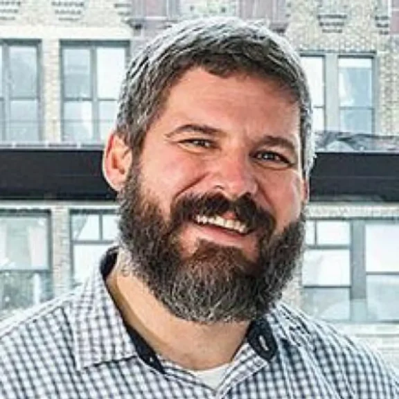 A man with a full beard and short hair, wearing a plaid shirt, smiles in front of large windows with a brick building in the background.