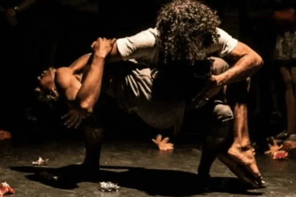 Two people perform a contemporary dance pose on a dimly lit stage, with one person balancing horizontally across the other's bent legs. Scattered objects or decorations are on the floor around them.