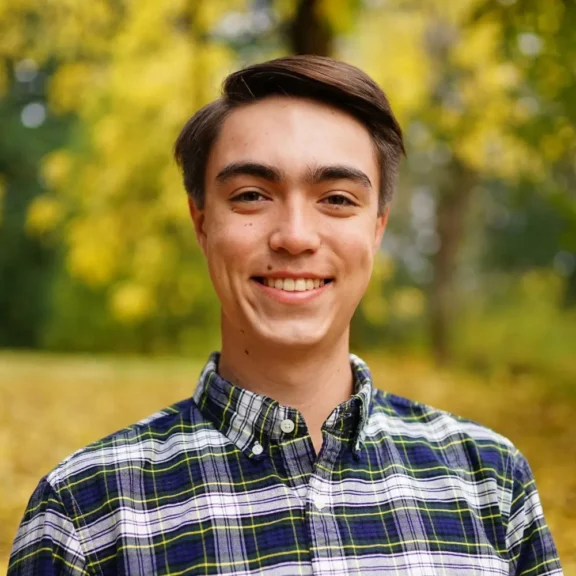 A young man with short brown hair wearing a plaid shirt, standing outdoors in a park with yellow autumn foliage in the background.