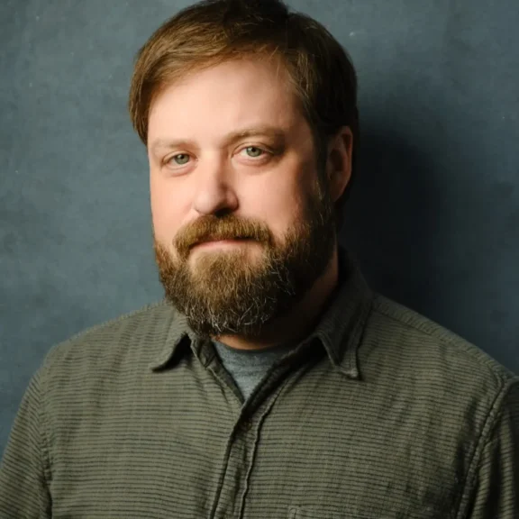 A man with light brown hair and a beard is wearing a textured button-up shirt and looking at the camera against a plain blue-grey background.