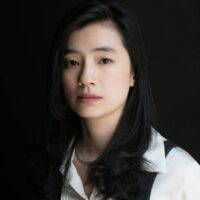 A woman with long black hair wearing a white blouse and black vest looks at the camera against a dark background.