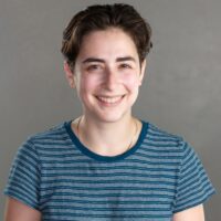 A person with short brown hair wearing a blue striped t-shirt is smiling at the camera against a plain gray background.