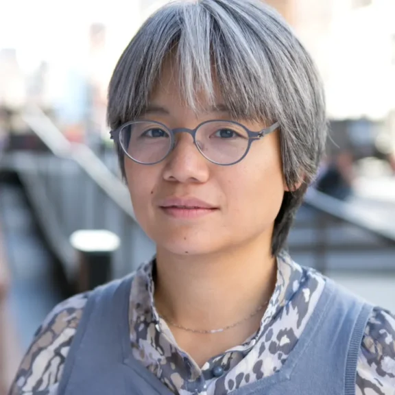 Person with short gray hair and round glasses wearing a patterned shirt and gray vest, standing outdoors in an urban setting.