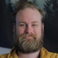 A man with a full beard and mustache, wearing a mustard-colored shirt, smiles at the camera with a blurred forest scene in the background.