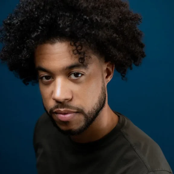 A man with curly hair and a trimmed beard looks at the camera against a solid blue background, wearing a dark shirt.