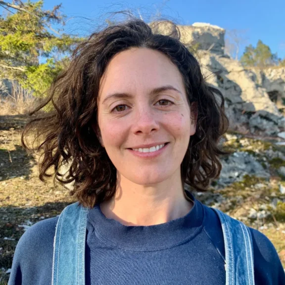 A person with curly brown hair, wearing a blue sweatshirt and denim overalls, stands outdoors with rocks and trees in the background on a sunny day.