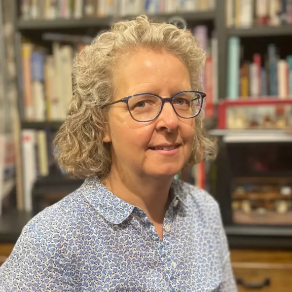 A woman with curly gray hair and glasses, wearing a patterned shirt, stands in front of a bookshelf filled with books and various items.