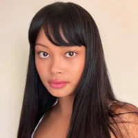 A young woman with long straight black hair and bangs looks directly at the camera against a plain light background.