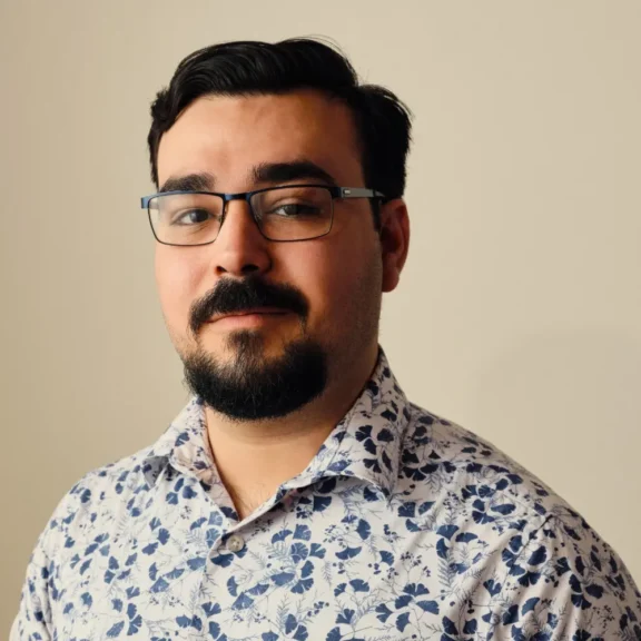 A man with short dark hair, glasses, and a beard is wearing a white shirt with a blue floral pattern, standing in front of a plain beige background.