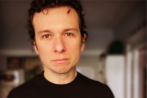 A man with short curly dark hair and a neutral expression stands indoors, wearing a black shirt. The background is softly blurred.