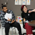 A man holding papers and a woman with a pencil sit together at a table in a classroom, both looking toward the same direction and smiling.