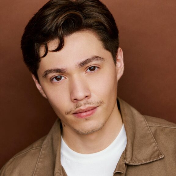 A young man with short brown hair and a light mustache looks at the camera, wearing a tan jacket over a white shirt against a brown background.