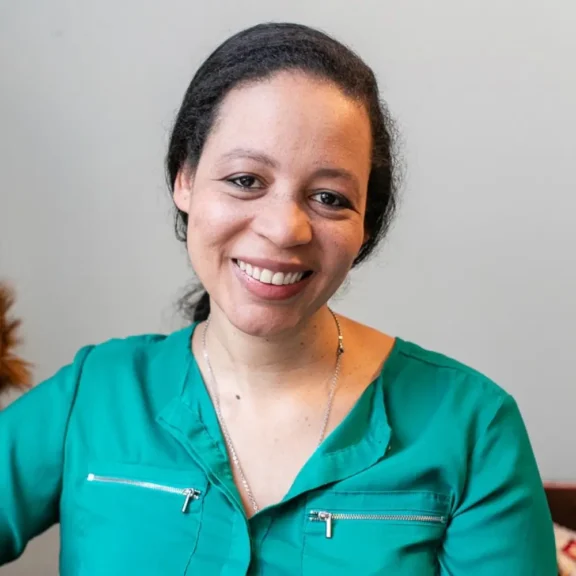 A woman with medium skin tone and dark hair smiles at the camera, wearing a green zippered blouse and a silver necklace, seated indoors against a plain light background.