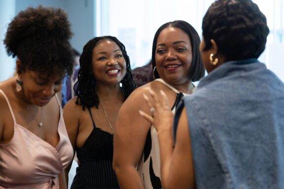 Four women stand together indoors, smiling and conversing in a well-lit room. Two are facing the camera, while two are turned slightly away.