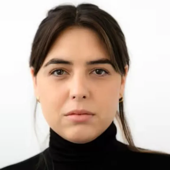 A woman with straight brown hair, parted in the middle, wearing a black turtleneck and small gold hoop earrings, looks directly at the camera with a neutral expression.