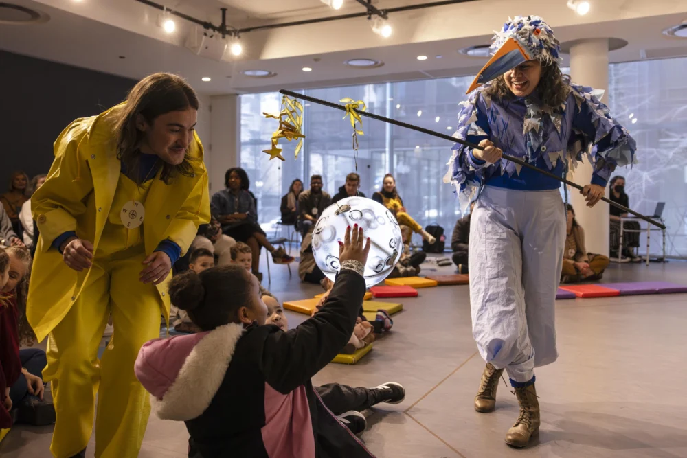 Two performers in colorful costumes interact with a seated child, who reaches for a glowing prop, while an audience watches in a well-lit indoor space.