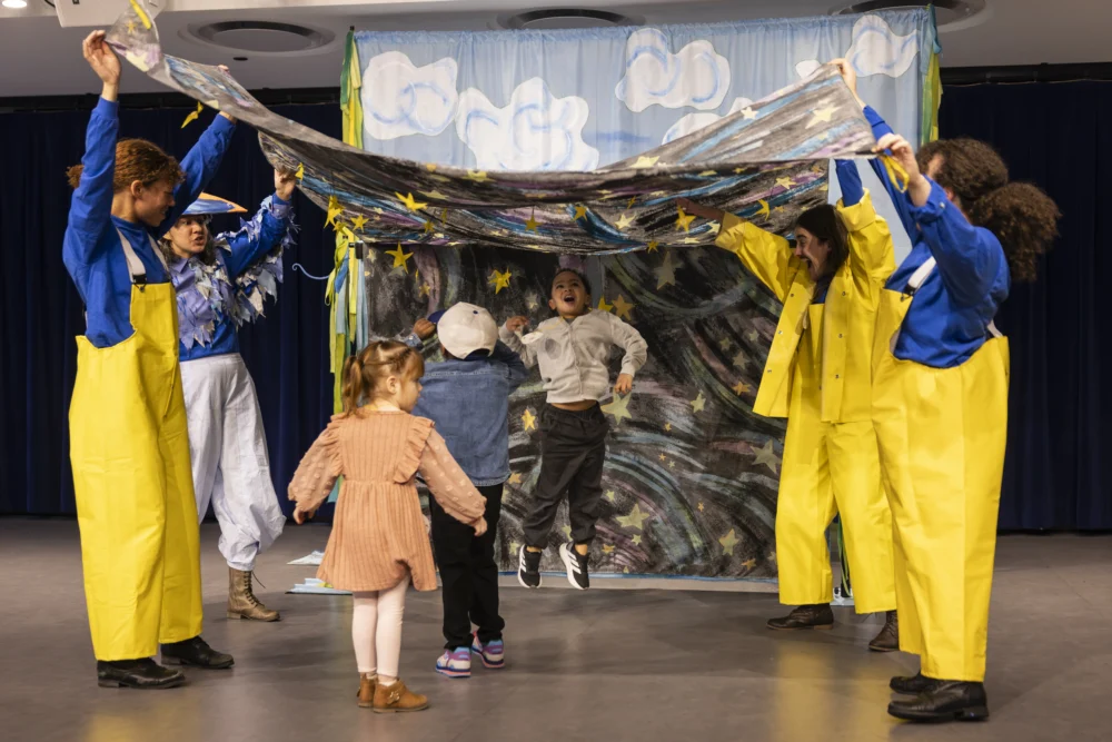 Several adults in costumes hold up a painted sheet while children walk and jump underneath it on a stage decorated with clouds and stars.