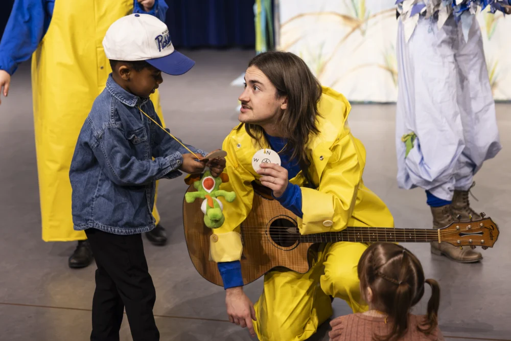 An adult in a yellow raincoat kneels with a guitar, showing a circular object to a child in a denim jacket and cap; another child sits nearby.