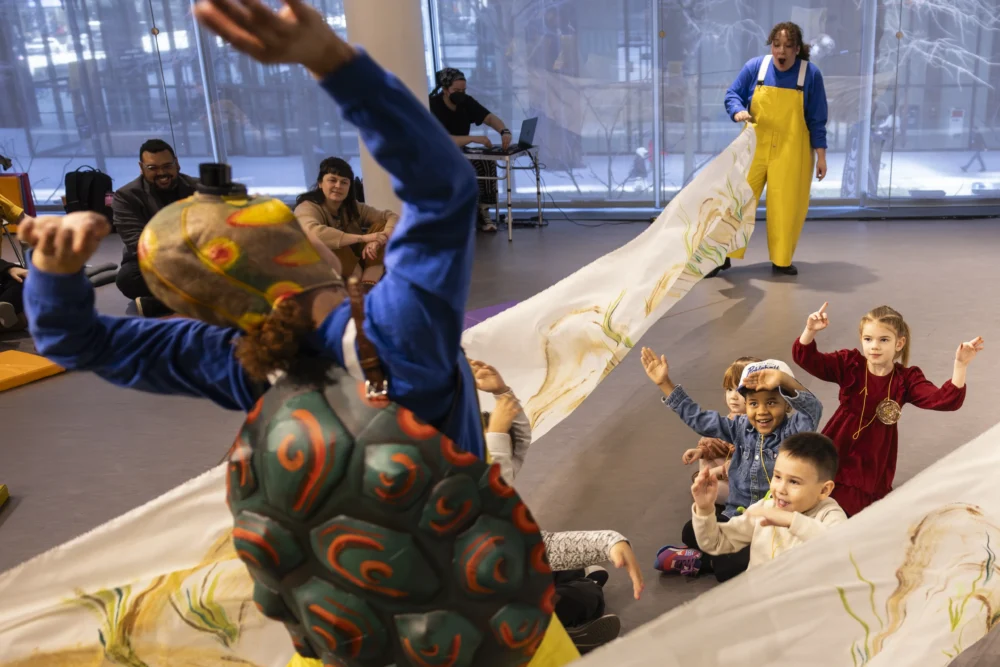 A performer in a turtle costume leads a group of children seated on the floor, while another adult holds a painted fabric backdrop indoors.