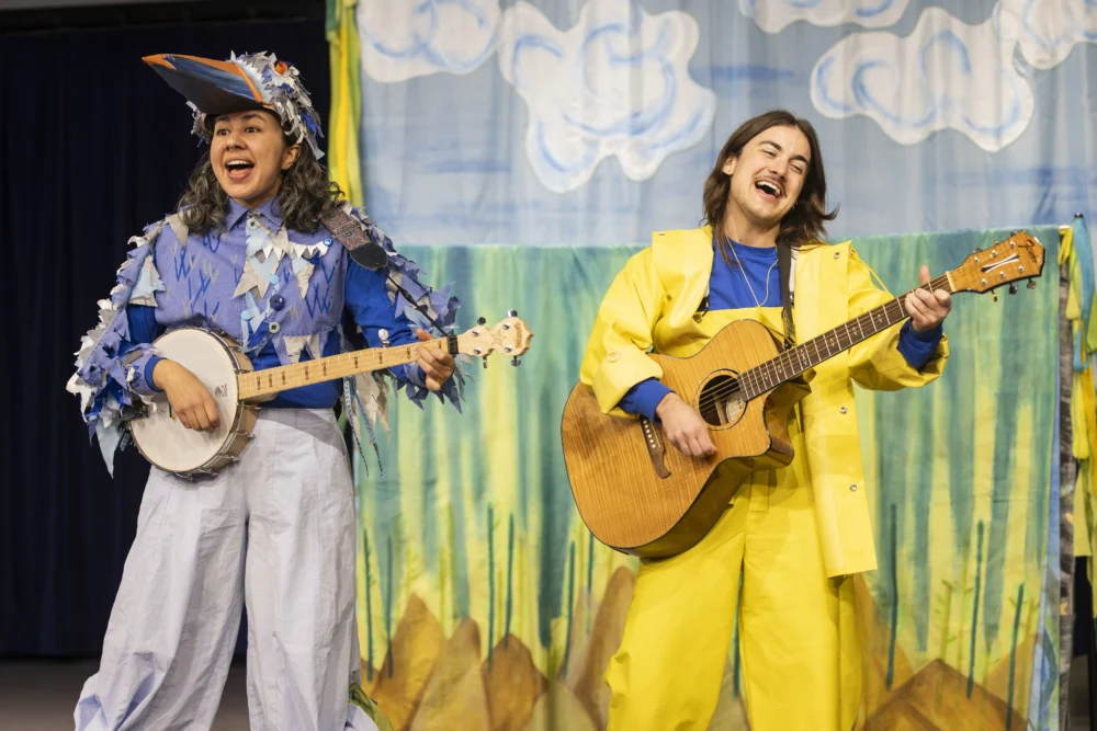 Two performers on stage: one dressed as a bird playing a banjo, the other in a yellow raincoat playing an acoustic guitar, with a painted backdrop behind them.