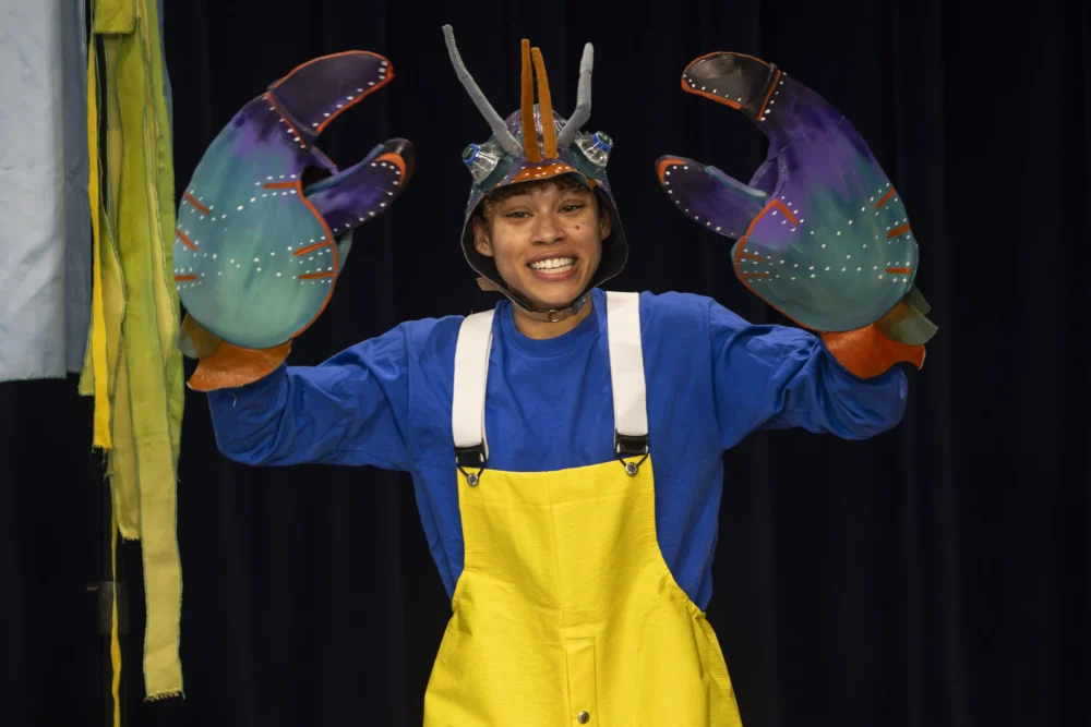 Person in a blue shirt, yellow overalls, and a helmet with antennae poses with large, colorful lobster claw gloves against a dark background.