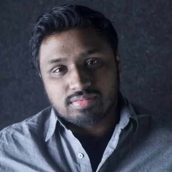 A man with short dark hair, a beard, and a light gray button-up shirt looks at the camera against a dark background.