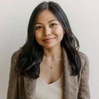 A woman with long dark hair wearing a brown blazer and a cream top stands against a plain light background, smiling slightly at the camera.