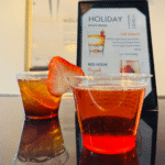 Two plastic cups with cocktails sit on a table; one is red with a strawberry slice garnish, and the other is amber-colored. A drink menu stands in the background.