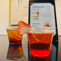 Two plastic cups with cocktails sit on a table; one is red with a strawberry slice garnish, and the other is amber-colored. A drink menu stands in the background.