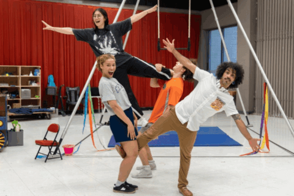 Four people practice a group acrobatic pose in a gym-like setting with mats, props, and a red curtain in the background.