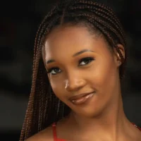 A woman with braided hair and a slight smile looks at the camera against a dark background.