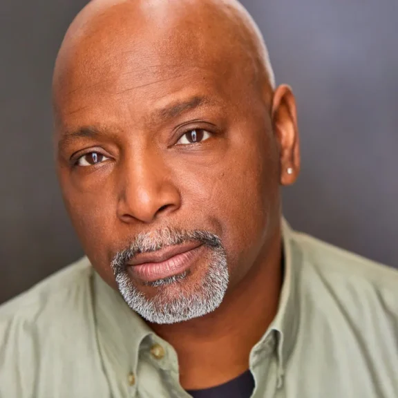 A middle-aged man with a shaved head, trimmed gray beard, and mustache, wearing a light green collared shirt, looks directly at the camera against a plain background.