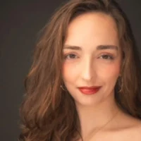 A woman with long wavy brown hair, light skin, and red lipstick is facing the camera against a dark background.