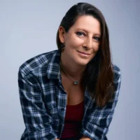 A woman with long brown hair, wearing a plaid shirt over a red top, sits and looks at the camera against a plain background.