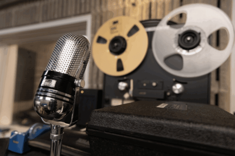 A vintage microphone is positioned in front of a reel-to-reel tape recorder in a recording studio setting.